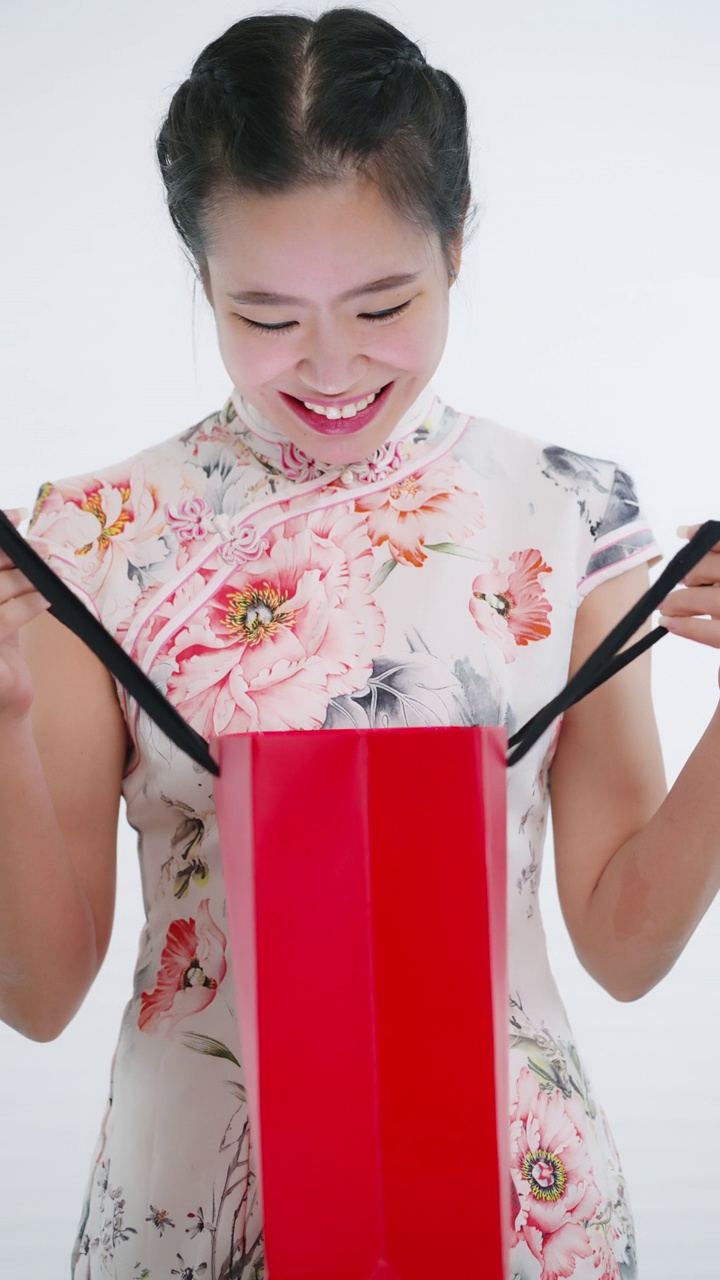 中国/亚洲妇女在春节期间打开红色购物袋迎接一个惊喜，白色的背景，穿着旗袍白色花卉图案的衣服视频下载