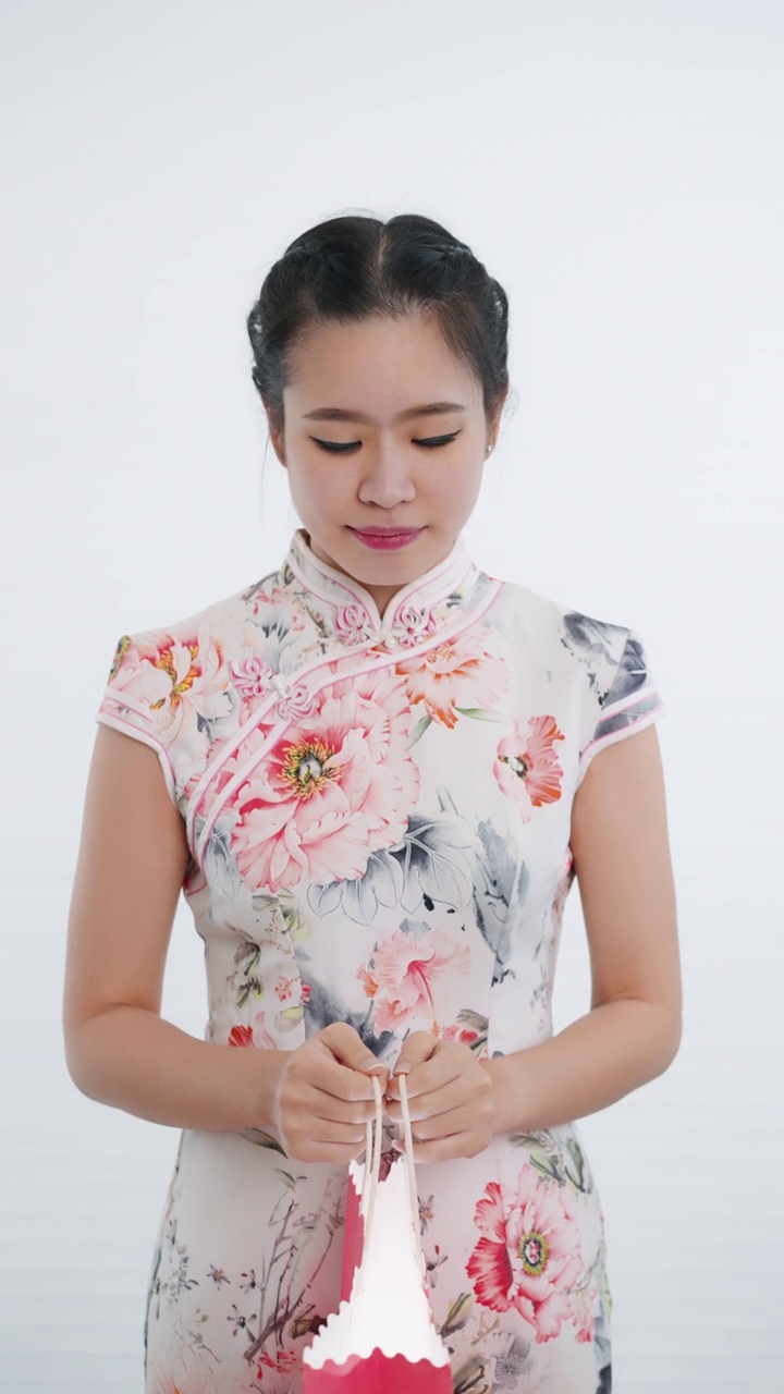 中国/亚洲妇女在春节期间打开粉红色的购物袋，在白色的背景下，穿着旗袍白色花卉图案的衣服，遇到了一个惊喜视频下载