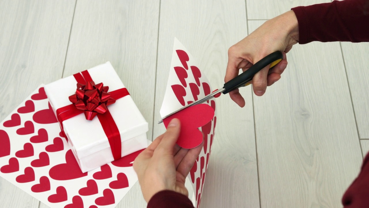 一名女子用剪刀在纸板上剪出红心的形状。DIY问候心礼物送给妈妈或爱人关怀。手工制作的情人节装饰和礼盒。爱的概念。视频下载