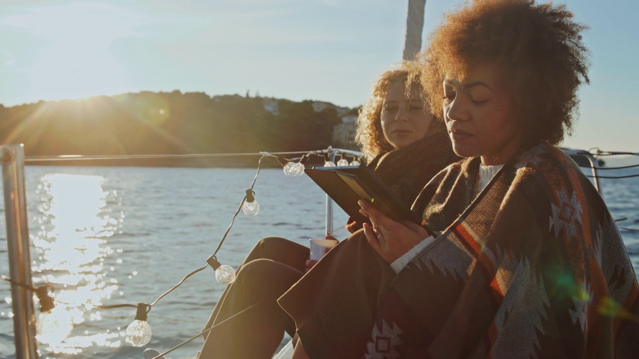 日出时分，在一艘帆船的甲板上，两名年轻女性在使用数码平板电脑时感到寒冷视频素材