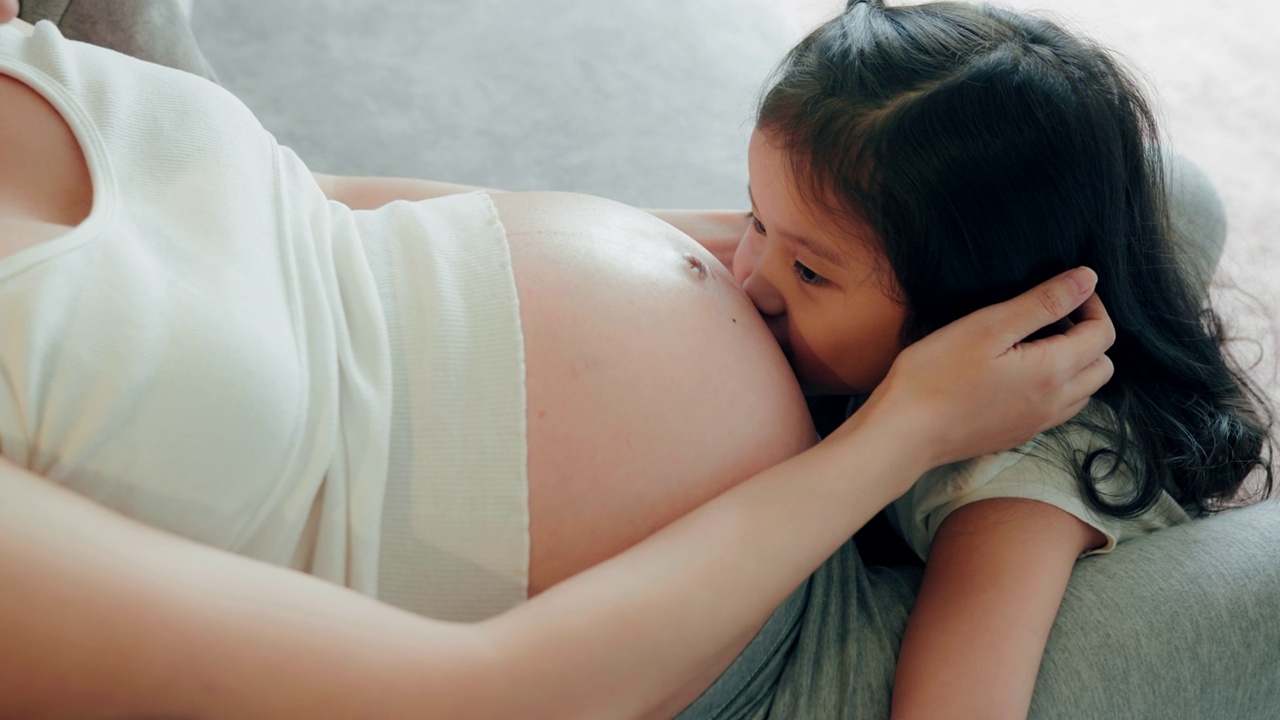 怀孕的母亲和学龄前的女儿坐在家里的沙发上。可爱的女孩亲吻怀孕妈妈的肚子。视频下载