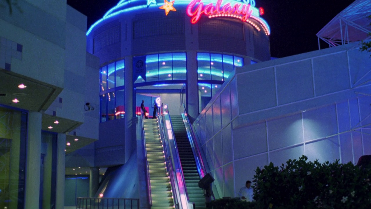 从好莱坞大道向右向左平移。好莱坞银河购物中心的霓虹灯招牌“好莱坞银河”。街上停着汽车，人们走在好莱坞星光大道上。街灯一字排开。在楼梯和自动扶梯上的人。里面有霓虹灯视频下载