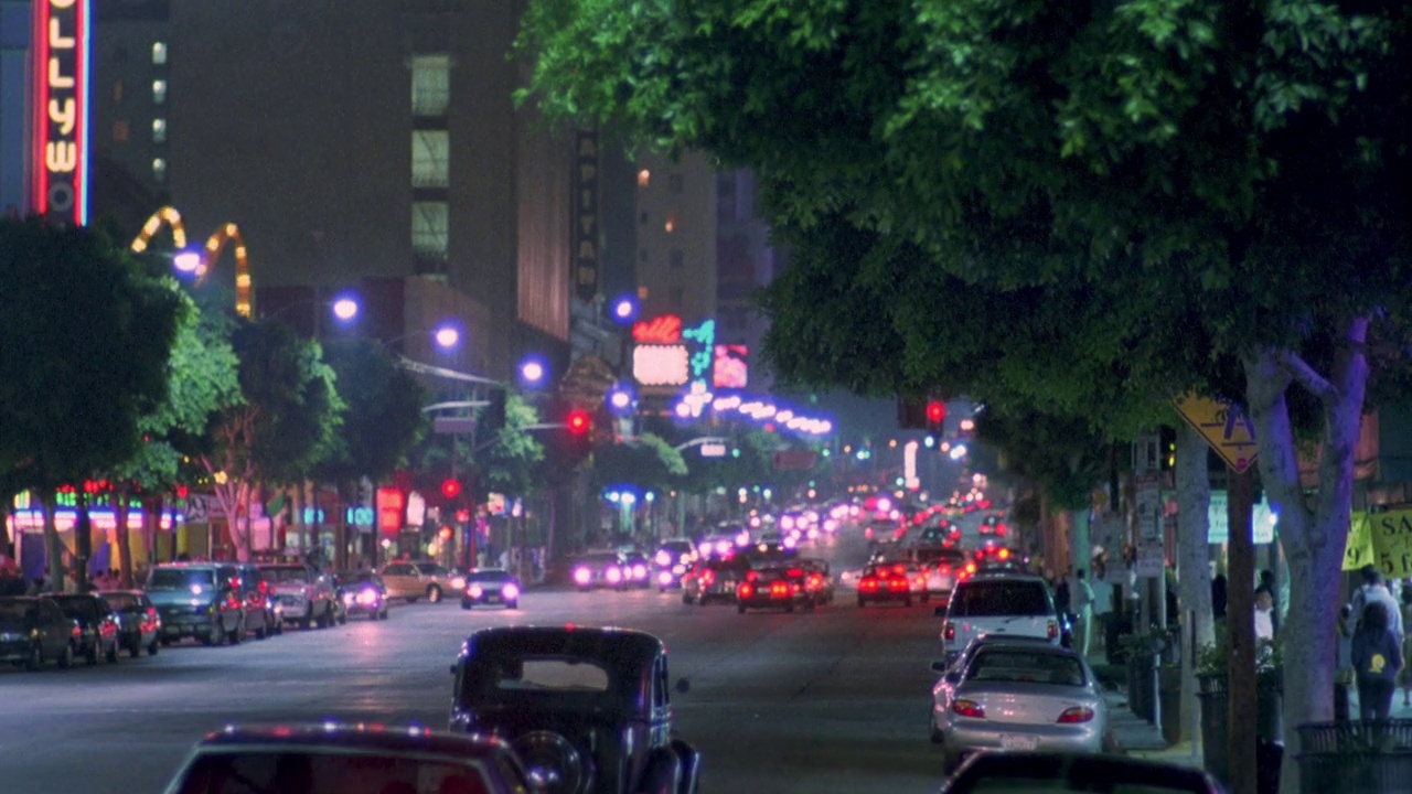 从城市街道到好莱坞大道上的汽车。霓虹灯、树木和商店排列在街道两旁。汽车驾驶。车辆停放。行人走在人行道上。视频下载