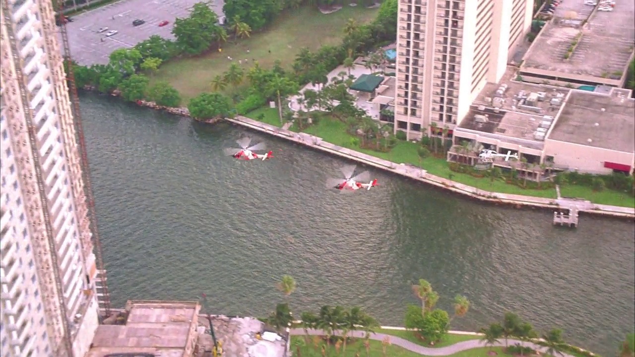 两架橙色和白色的海岸警卫队直升机和一架白色的警察直升机在迈阿密的海岸线或沿海水域上空飞行。直升飞机排成单行在高层公寓楼后面飞行。视频素材