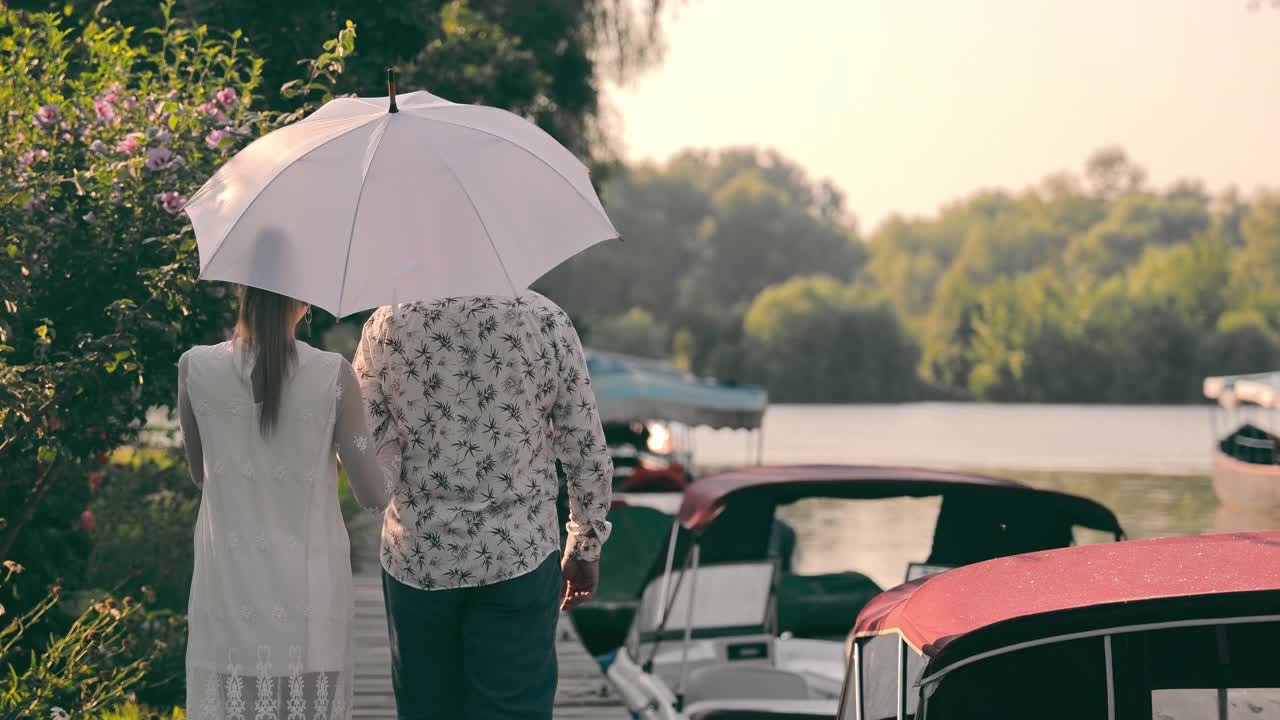 一个男人和一个女人沿着长廊散步并交流。一个男人在太阳下撑着一把白色的伞。一对年轻的夫妇走在摩托艇码头附近视频下载