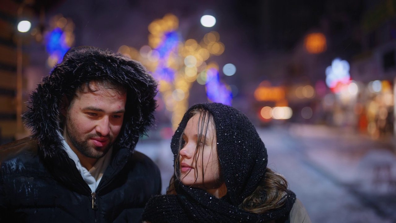年轻的情侣约会，在街上的咖啡馆喝热饮暖身，在城市的夜晚下雪视频下载