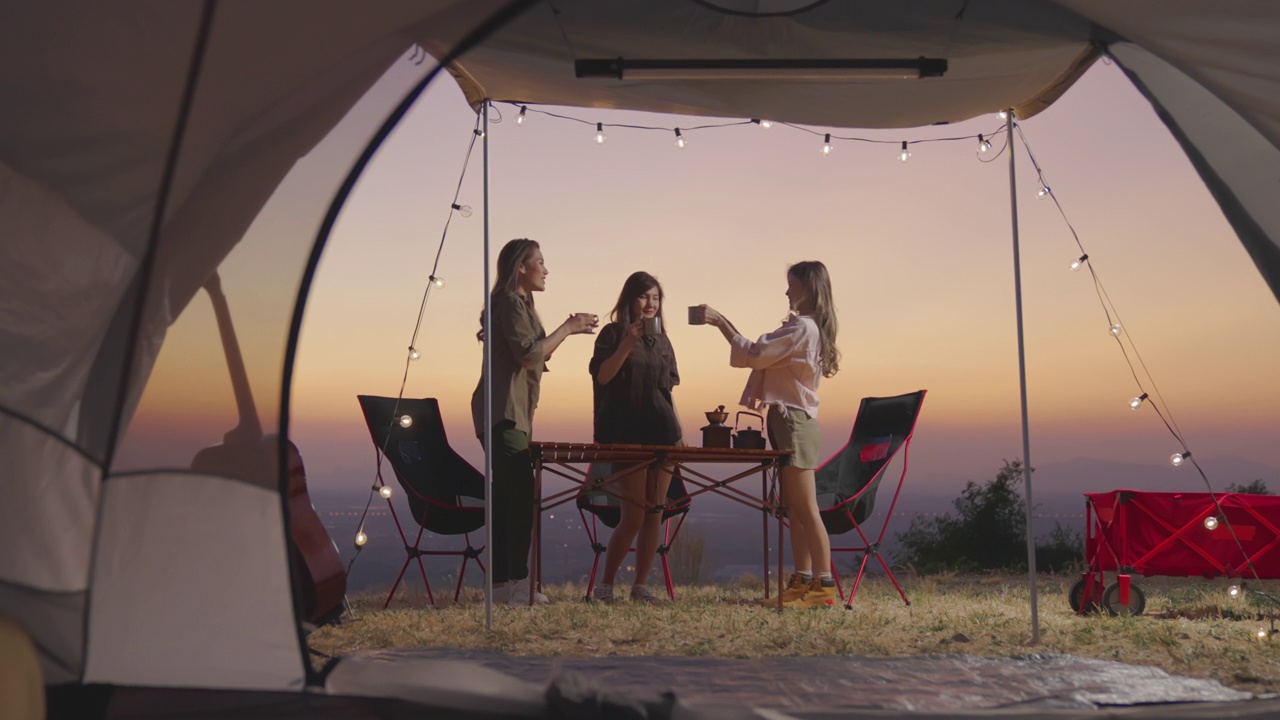年轻女子休息在旅游帐篷和燃烧篝火看日落与你的朋友在假期。亚洲人，野营，朋友，帐篷，日落，游客。视频下载