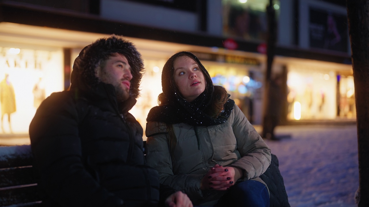 年轻的情侣约会和坐在长椅上聊天，而在城市的雪晚上视频素材