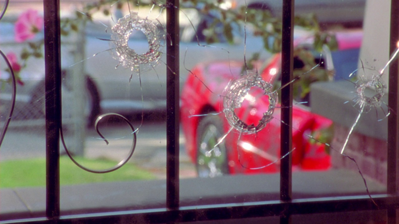 房子或公寓窗户的中角，前面有栅栏。窗户有一个洞。部分窗户呈圆形破碎表明是被枪击或子弹击中。可能被用于帮派暴力。看到银车和红车停在车道上。看到两个铁道部视频下载