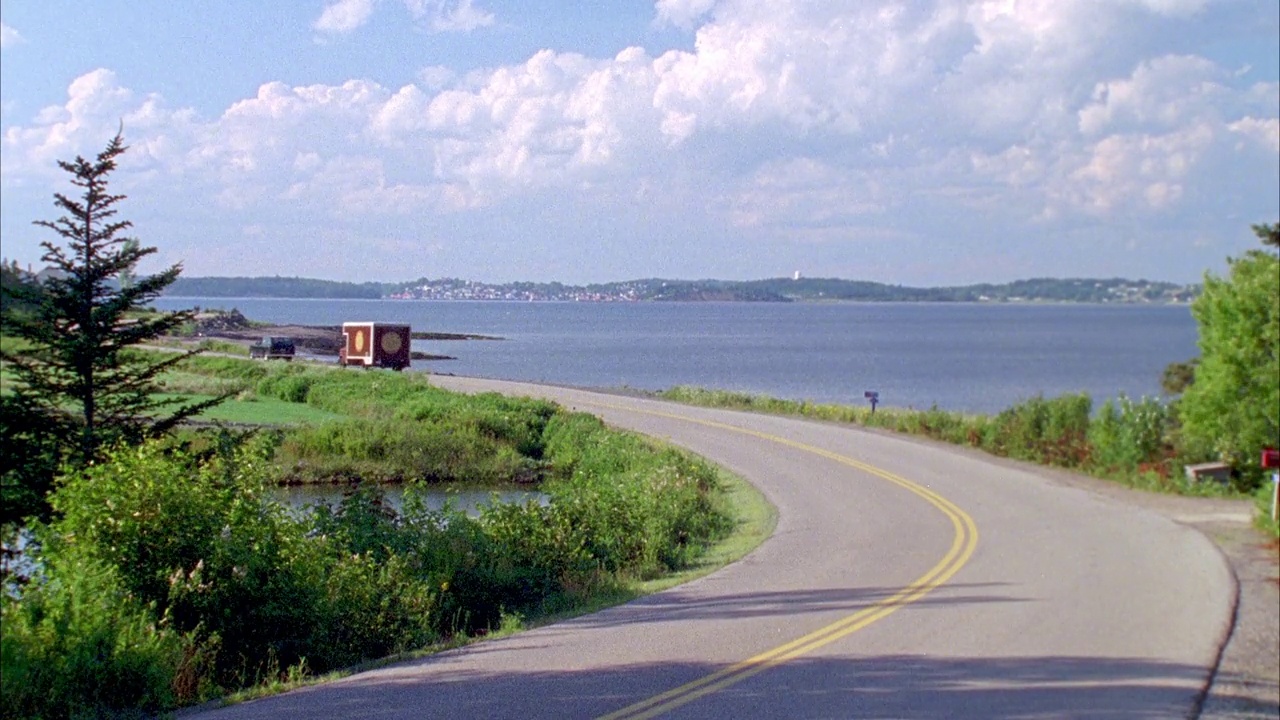 广角两车道的海滨或湖边路。见送货卡车追逐小型吉普车。在背景中可以看到灯塔、山丘、小镇和水。可能是飞车追逐。视频素材
