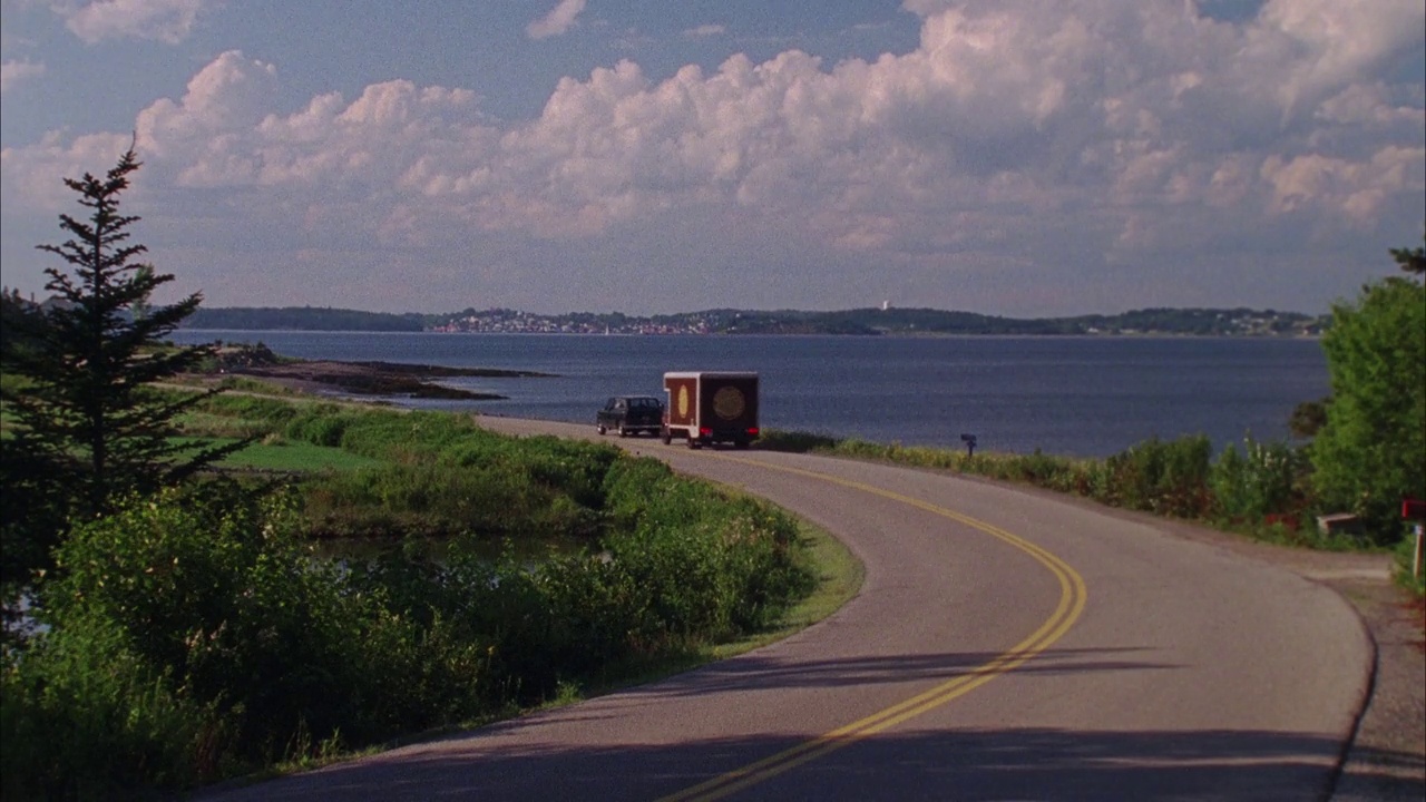 广角两车道的海滨或湖边路。见送货卡车追逐小型吉普车。在背景中可以看到灯塔、山丘、小镇和水。可能是飞车追逐。视频素材