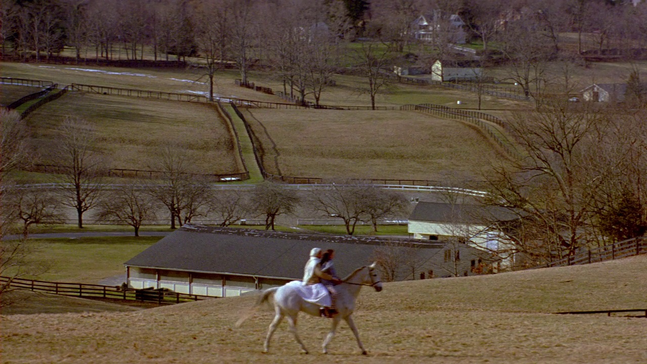 广角的灰色天空，山脉和树木，向下平移到农场地区的小山。两个女人骑马穿过田野。视频下载