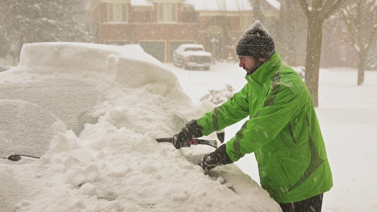 一场暴风雪中，一名年轻的成年男子正在清理车上的雪。视频下载