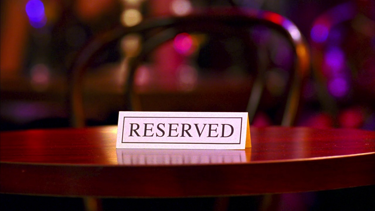 酒吧或夜总会预定餐桌的特写镜头。一只手拿起白色卡片，上面写着“保留”。背景色灯。人们走过桌子。架的焦点。视频下载