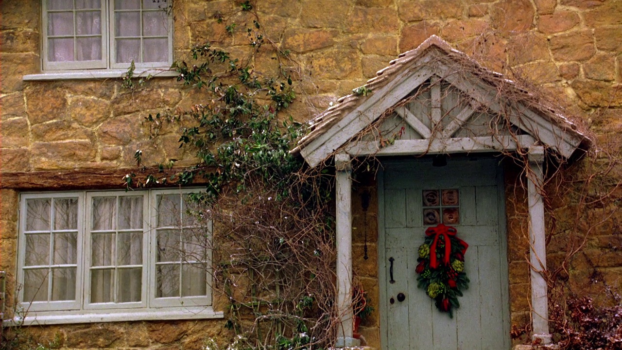 中角两层砖或石砌的村舍或房屋。一层和二层的白色平开窗。绿色的常春藤长在柱子上和前门的山墙上。节日或圣诞花环挂在门上。山墙上有些雪。冬天。视频下载