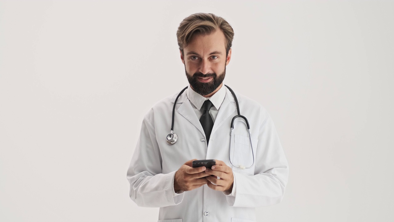 带听诊器的帅哥医生一边用智能手机一边微笑眨眼视频素材