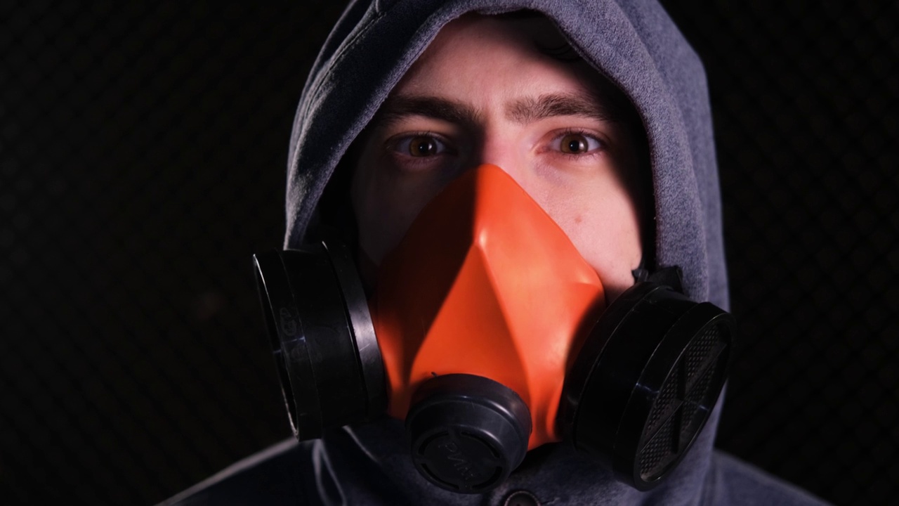 一个戴着兜帽和橙色呼吸器的男人抬起头，在黑暗中看着摄像机。防护面具。环境污染的概念视频下载