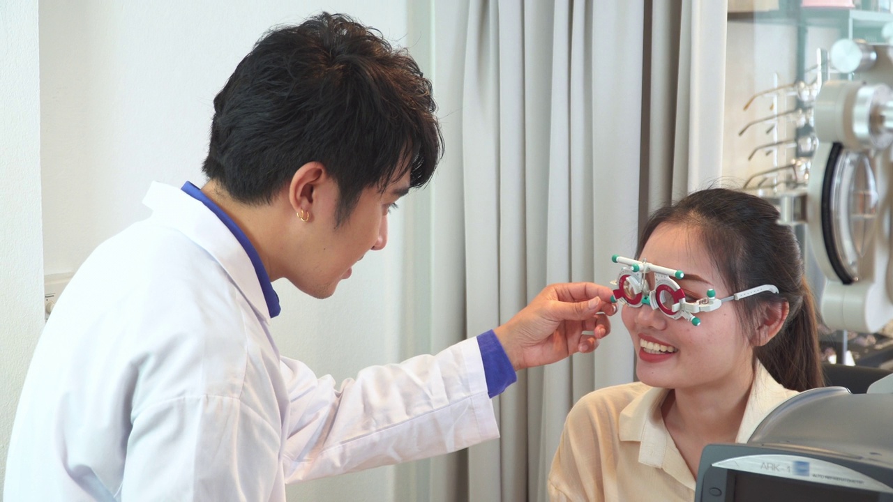 验光师在插入镜片后调整距离试镜架，测试亚洲年轻女性的视力，阅读视力表，Snellen视力表，准备制作眼镜，表达积极的情绪，微笑以获得更好的视力视频下载