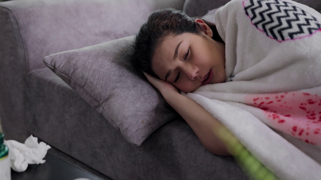 近景:发高烧的亚洲病弱母亲躺在沙发上，感到强烈的头痛，盖着毯子，在家擦鼻子。疾病季节性病毒问题感冒和偏头痛。视频下载