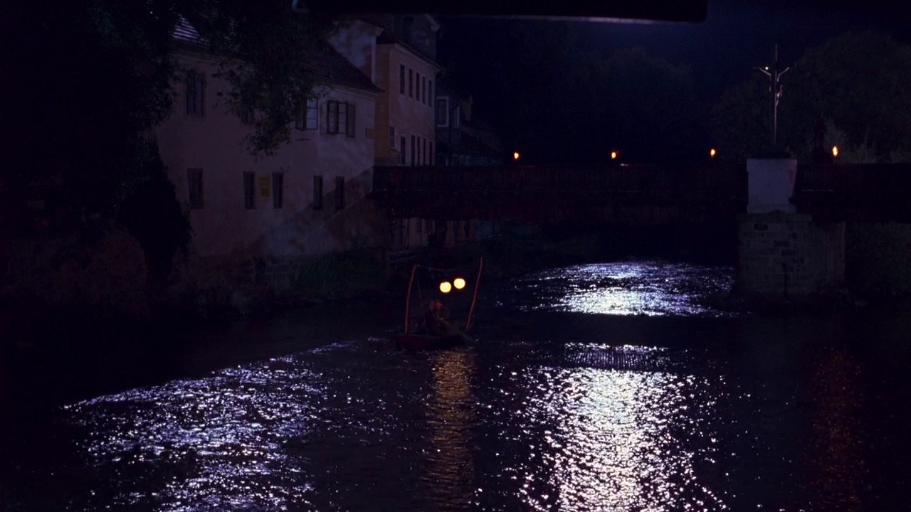 沿运河顺流而下的小船或划艇的广角。可能是一条河。水上的一座小桥连接着一座多层住宅，也许是一栋公寓楼。用发光的灯装饰的船，像节日用的灯。欧洲。视频下载
