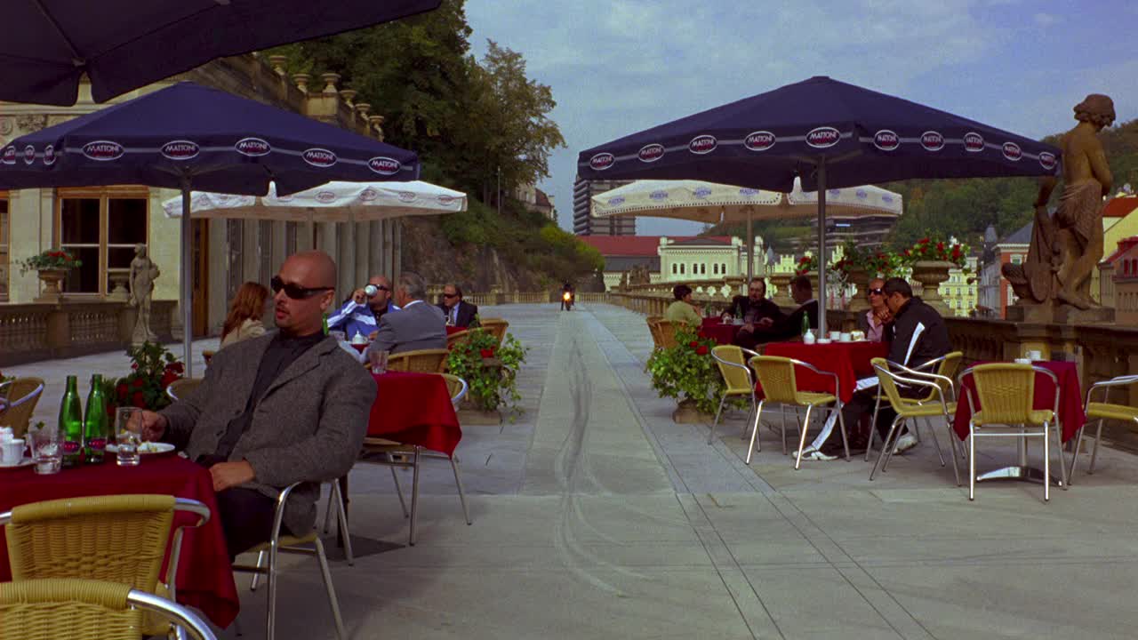 欧洲城市咖啡馆或餐馆的广角拍摄。一个穿夹克戴太阳镜的男人坐在桌子旁。一个骑摩托车的人从桌子旁开过，从后面拿出一个纸板箱，把它带到镜头前。快递或快递员的视角。的起源视频下载
