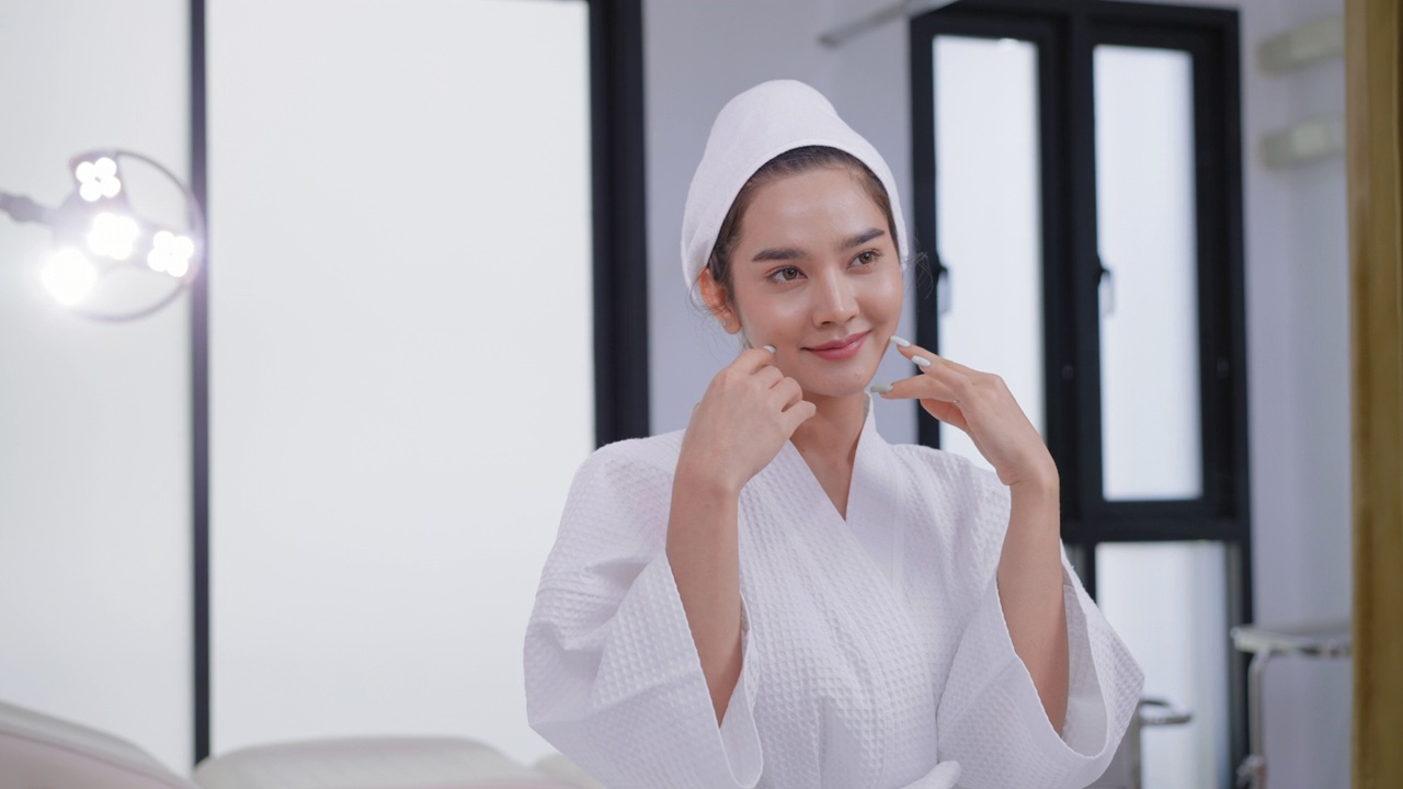 快乐可爱的亚洲妇女在白色浴衣举起手臂摆姿势在美容诊所的肖像。年轻美丽的亚洲妇女在浴衣和毛巾包裹头部微笑与自然健康的皮肤。视频素材