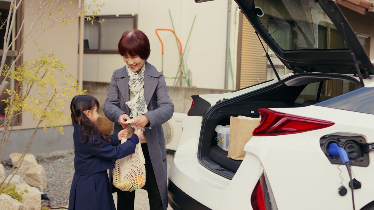 MS-一个年轻的女孩正在帮助她的祖母从他们的电动汽车上卸下购物的东西视频下载