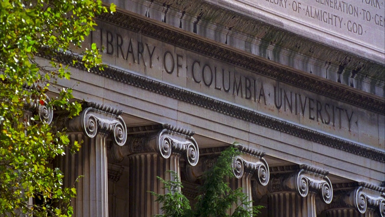 从巴特勒图书馆的浮雕往下看，上面用离子柱写着“哥伦比亚大学图书馆”。一名女大学生坐在台阶上，另一名学生坐在站台上。视频下载