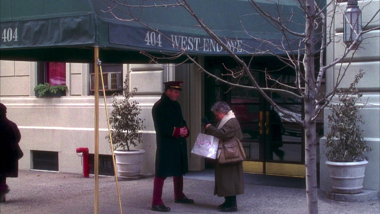公寓大楼入口和雨篷的中角，雨篷上写着“404 west end ave.”老妇人向看门人展示了一个购物袋和一个盒子，他们一起走进了门。摇到三楼的窗户上。视频下载