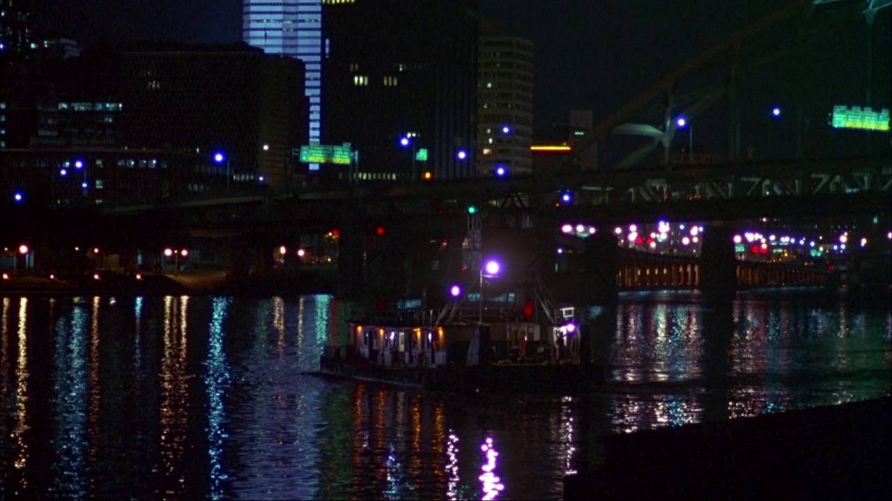 船的角度适中，可能是船屋，在河流或水边漂流。桥下渡船，背景是市中心。城市的灯光反射在水面上。视频下载