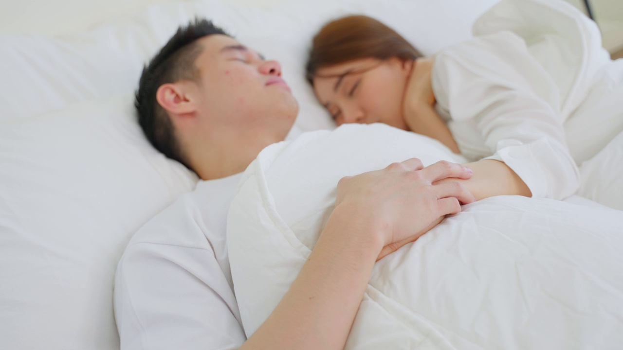 亚洲有吸引力的年轻夫妇躺在床上睡觉在卧室。美丽的新婚夫妇，穿着睡衣的男人和女人，清晨在房子里一起躺在舒适的枕头上，感到幸福和放松。视频素材