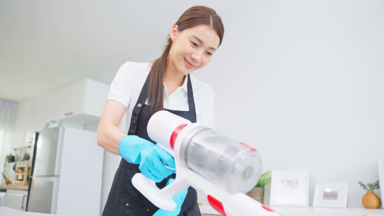 亚洲清洁服务女工清洁客厅在家里。美丽快乐的女孩，家庭主妇管家清洁工穿围裙和吸尘凌乱肮脏的桌子家务或家务。视频素材