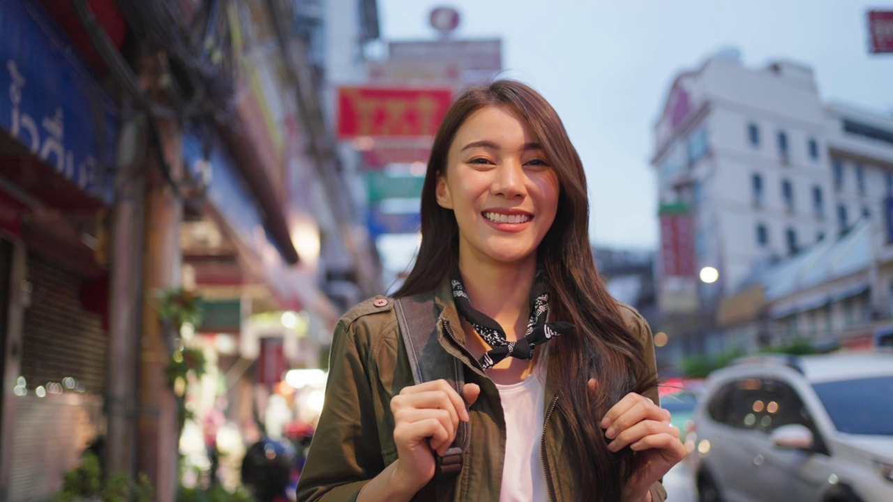 亚洲有吸引力的女人背包客旅行的肖像在城市的夜晚。年轻的女游客站在美丽的街道享受花时间度假旅行在泰国，看着相机。视频素材
