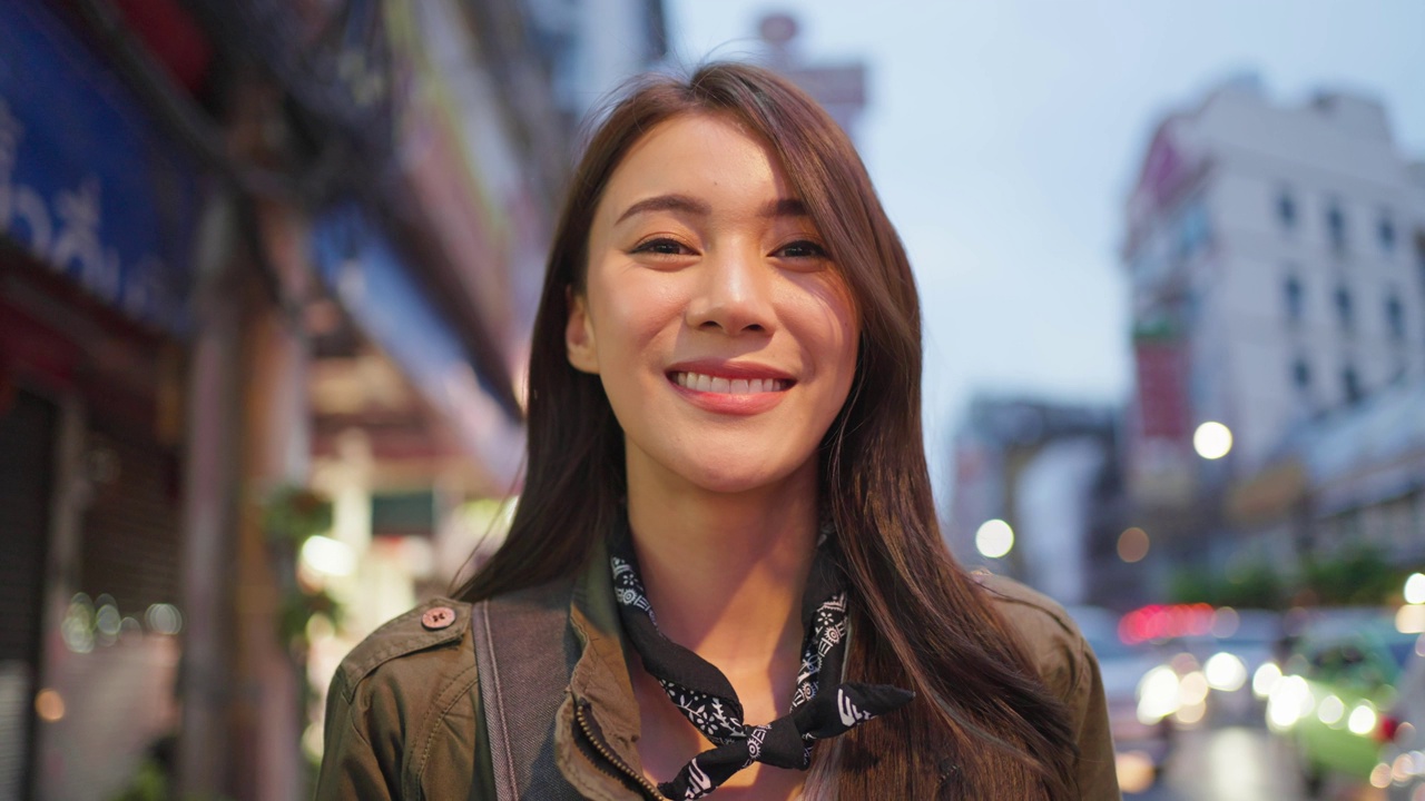 亚洲有吸引力的女人背包客旅行的肖像在城市的夜晚。年轻的女游客站在美丽的街道享受花时间度假旅行在泰国，看着相机。视频下载
