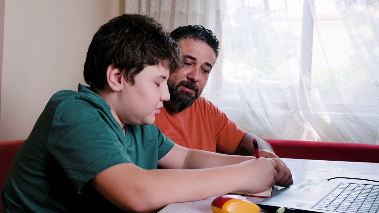 愤怒的父亲帮助压力大的儿子在家做作业，而坐在桌子前的笔记本电脑视频下载