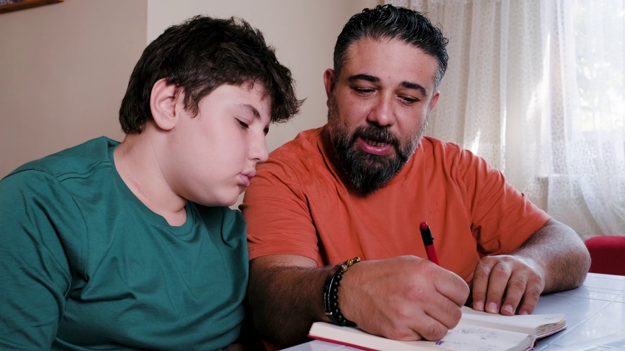 愤怒的父亲帮助压力大的儿子在家做作业，而坐在桌子前的笔记本电脑视频下载