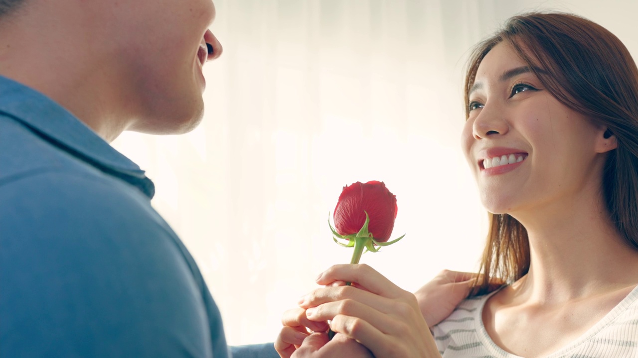 亚洲青年惊喜送玫瑰给美丽的女友。迷人浪漫的新婚男女花时间在房子里一起庆祝周年纪念日和情人节。视频下载