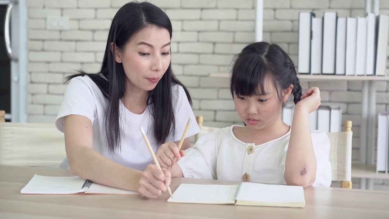 精疲力竭的年轻亚洲单身母亲头痛，教小女儿做作业。小女孩不懂家庭作业，她妈妈对此很不高兴。孩子的教育问题。视频下载