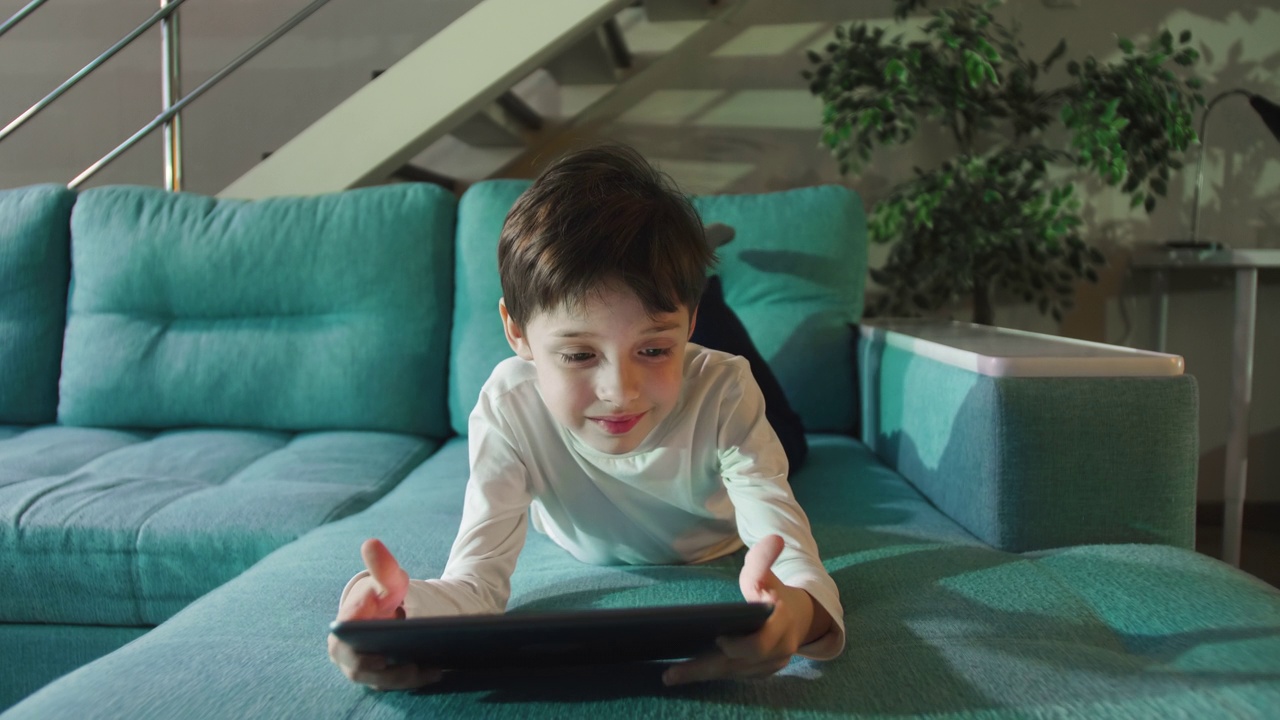 一个小男孩独自躺在沙发上，使用数码平板电脑。视频素材