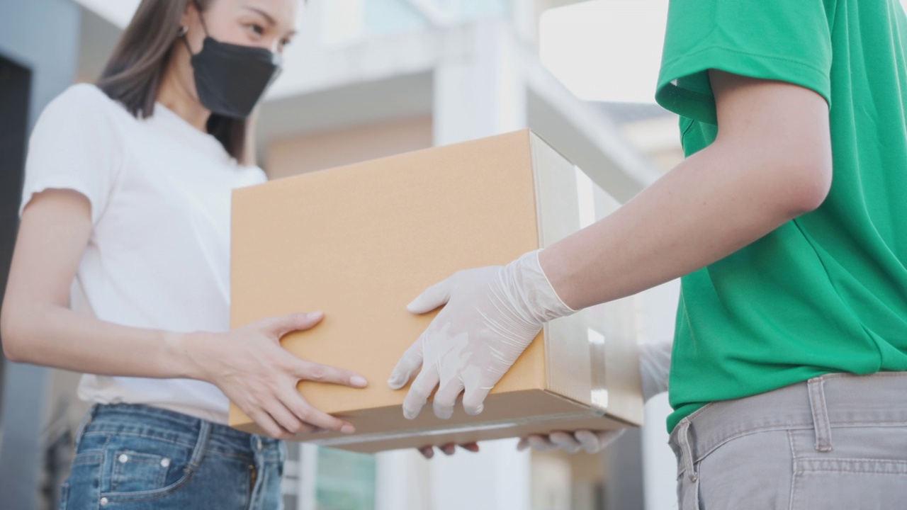 中景镜头。亚洲快递员穿着绿色制服，戴着防护口罩和医用橡胶手套，在客户家门口送一个纸箱给客户。4 k决议。视频素材