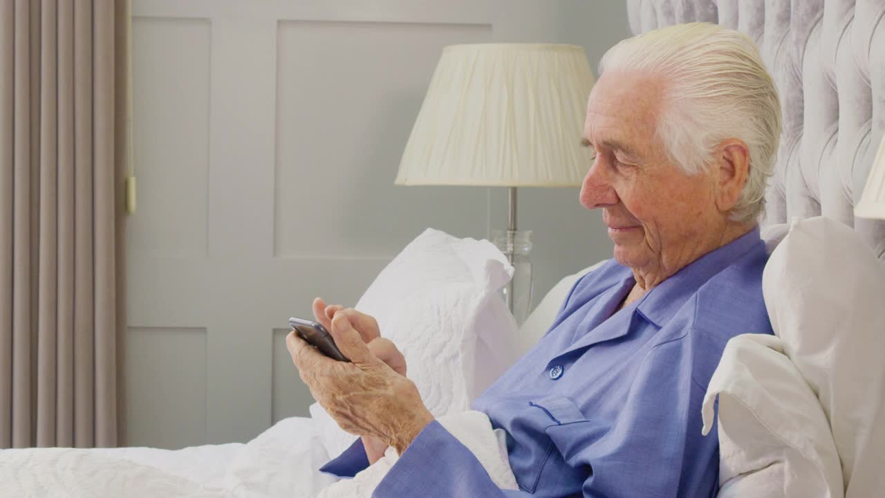 微笑的老人在家穿着睡衣在床上用手机视频素材