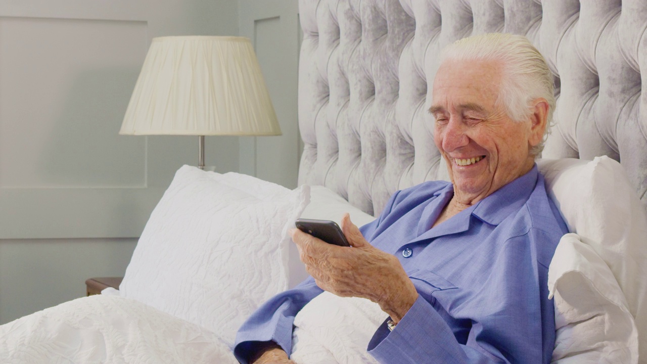 老人在家里穿着睡衣在床上做手机视频通话视频素材