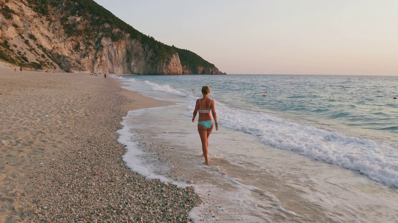 空中低空跟随成年妇女享受美丽的米洛斯海滩的Lefkada，爱奥尼亚岛，希腊。夕阳金色的阳光和原始的海浪打破了蓝绿色的大海在岸边视频下载
