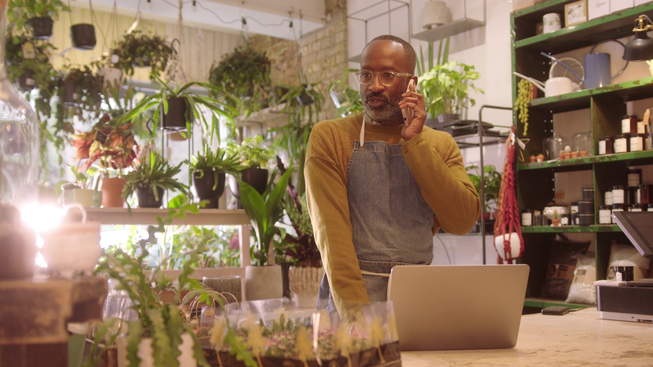 男性花商在植物商店用笔记本电脑工作，用智能手机聊天视频购买