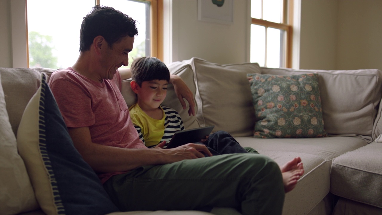 微笑的父亲看着儿子一起坐在客厅的沙发上玩平板电脑游戏的中宽镜头视频下载