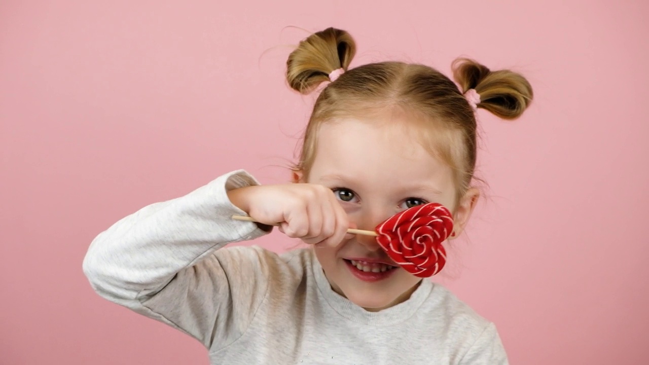 有趣的金发小女孩微笑着，在粉红色的背景上玩红心形状的棒棒糖焦糖。情人节或母亲节的概念视频下载
