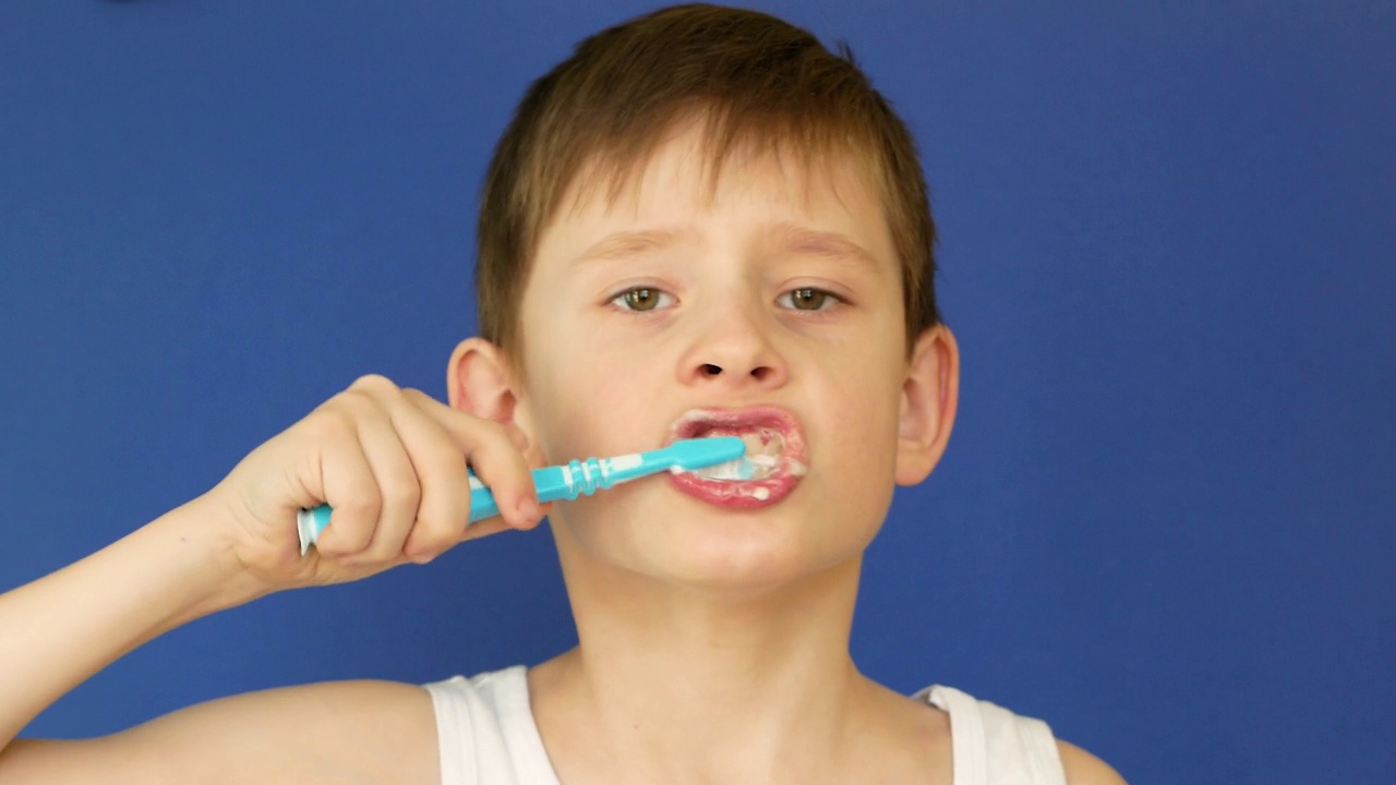 一个7岁的白人男孩在蓝色背景上刷牙的肖像。工作室拍摄。这孩子早上洗脸。儿童的牙齿卫生。预防儿童蛀牙。视频下载