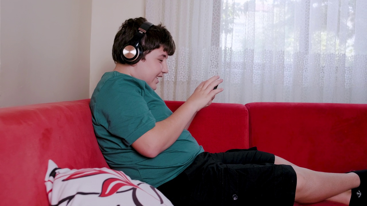 男孩在客厅用耳机玩多人在线游戏，使用智能手机连接视频素材