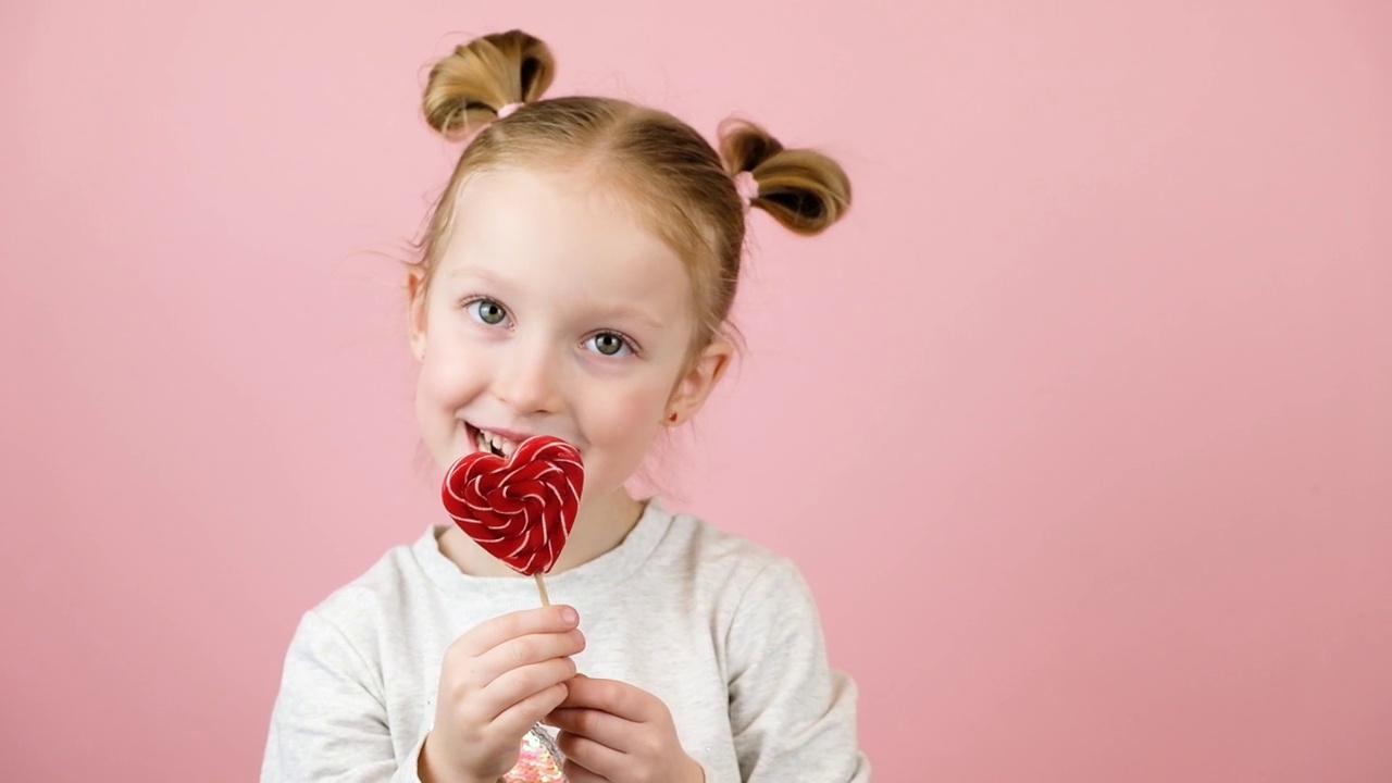有趣的金发小女孩微笑着，在粉红色的背景上舔着红心形状的棒棒糖焦糖。情人节或母亲节的概念视频下载