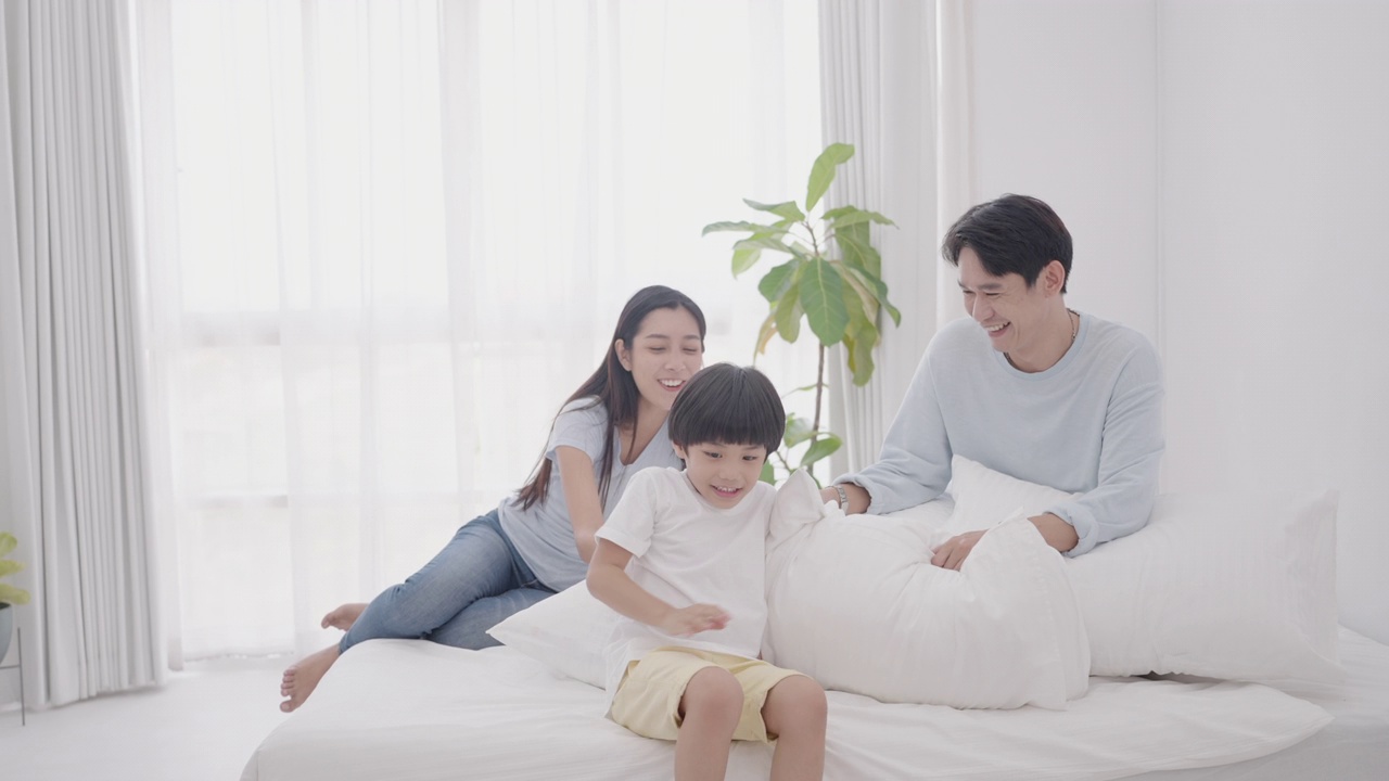 亚洲幸福家庭爸爸，妈妈和可爱的儿子，在舒适的家里享受周末。爸爸和妈妈一起坐在舒适的床上，微笑着看着儿子抱着枕头跳来跳去，玩得很开心。视频素材